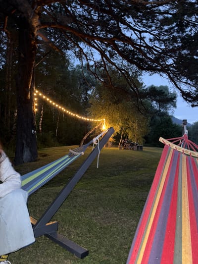 This media file captures a tranquil evening scene at Ust-Sema, Russia, with two hammocks set up in a grassy area adjacent to a wooded landscape. The time of day appears to be dusk or early night, as indicated by the dim natural light and the illuminated string of warm-toned lights stretching across the background. One hammock, with red, yellow, and purple stripes, is suspended from a wooden beam and a dark metal frame. Another hammock, featuring blue and green stripes, is also supported by the dark metal frame.
The foreground is dominated by the dark, intricate branches of a large tree on the left, its silhouette stark against the twilight sky. In the middle ground, a line of small, glowing bulbs adds a festive and inviting ambiance, casting a soft light on the surrounding grass and trees. Beyond the hammocks, the landscape features a gently sloping lawn leading to a dense line of trees and further hills or mountains in the distance. The overall atmosphere is one of relaxation and serenity, suggesting a peaceful evening spent outdoors. No visible text is present in the image.