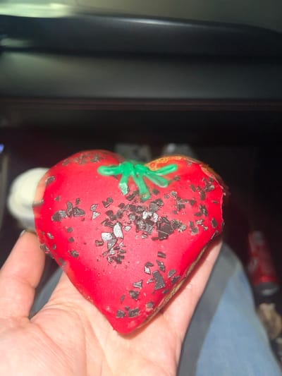 A partially eaten, heart-shaped donut sits in a hand. The donut is covered in red frosting and decorated with scattered dark chocolate flakes and a green, stem-like design. A bite has been taken from the top. The background is out of focus but suggests an indoor setting, with a dark cabinet and a glimpse of other items on a surface nearby. The overall lighting is dim.