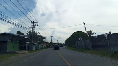 The image depicts a straight, asphalt road in Meunasahbaro, Indonesia, with a yellow line dividing the lanes. On the left side of the road, there are small, weathered buildings with corrugated metal roofs, one of which has green metal shutters. Power lines are strung across the sky, supported by numerous utility poles. Palm trees and other greenery are visible interspersed with the buildings. On the right side of the road, a concrete wall topped with a black metal fence runs along the property line. A dense canopy of trees borders the fence. A dark-colored SUV is driving away from the camera in the center of the road, and a few other vehicles are visible further down the road in the distance. The sky is partly cloudy with large white clouds against a pale blue backdrop. The scene appears to be an undeveloped or semi-developed area on the outskirts of a town.