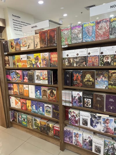 The image displays a well-stocked bookstore, likely a Gramedia branch, located in Dumai City, Indonesia, given the text "Gramedia" visible on a large sign. The shelves are filled with a variety of books, predominantly novels, arranged in neat rows.
The books cover a wide range of genres and authors. In the top section, there is a prominent display of "Harry Potter" books with their distinct cover art. Below, various other novels are shelved, including titles like "The Fifth Mountain," "Animal Farm," and works by Paulo Coelho and Gabriel Garcia Marquez. The second tier showcases more novels, with some featuring manga-style illustrations and titles such as "The Atala" and "The Girl Who Fell Beneath the Sea." The bottom shelves are also densely packed with books, including titles related to "Bungo Stray Dogs," "George Orwell's 1984," and other fiction.
The overall environment suggests a clean, organized, and professional retail space. The lighting is bright, likely from overhead store lighting, illuminating the colorful book covers. The flooring appears to be tiled. No people are visible in the frame, focusing the attention entirely on the merchandise. The arrangement and density of the books indicate a busy and popular bookstore.