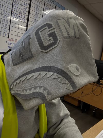 A person is depicted from a slightly elevated, oblique angle, seated in an indoor office environment in Nibley, United Kingdom. The individual's face is entirely obscured by the pulled-up hood of a grey hooded sweatshirt. The hood features raised appliqué designs, including the letters "N G M" in varying shades of grey across the upper portion, and below them, a distinctive shark-face motif with jagged teeth and a circular eye, rendered in similar grey tones. A neon yellow or chartreuse high-visibility garment, partially revealing a silver reflective stripe, is worn beneath the grey sweatshirt.

In the background to the left, a white magnetic whiteboard is mounted, displaying a grid filled with handwritten black numbers and text, including what appears to be inventory or financial data, and a partial quote. To the right, a section of an office workspace is visible, featuring a light brown wooden desk, a black computer monitor, and associated cables. The flooring is dark. The scene is illuminated by overhead indoor lighting.