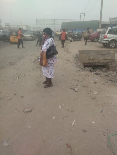 The scene is located in a dusty street in Way, Nigeria, at coordinates 9.029 latitude, 7.584 longitude. A woman, wearing a floral-print long-sleeved dress and a face mask, is standing on the side of the street, holding a brown shopping bag and a black handbag. Several other people are walking in the distance, and there are multiple cars parked on both sides of the street. The ground is dusty and uneven, with some debris scattered around. There are also a few power lines running overhead. The street is narrow and appears to be in a state of disrepair. The overall impression is one of a busy, bustling street in a developing country.