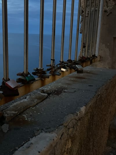 Metal bars run parallel against a hazy blue backdrop, secured to a weathered concrete wall. A row of padlocks, varying in size and color, are attached to the bars. Cracks and discoloration mar the wall's surface, indicating age and exposure. The scene is captured at an angle, emphasizing the texture of the concrete and the repetition of the bars.