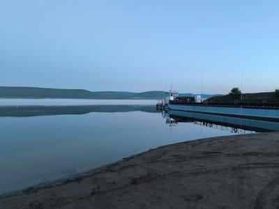 The image depicts a serene, pre-dawn or twilight scene on a body of water, likely a river, near the city of Gremuchiy, Russia. A long, flat-bottomed barge, painted a light blue, is moored along the right side of the frame. The barge has a small, white superstructure at its front, with visible railings and what appear to be communication masts. Life rings are attached to the railings. The water is calm, reflecting the barge and the hazy, low-lying mist that stretches across the horizon on the left side of the image. Beyond the mist, rolling hills are visible, silhouetted against the pale, clear sky, which transitions from a pale blue at the top to a lighter hue near the horizon. The foreground consists of a sandy or muddy bank, showing tire tracks, indicating recent activity. The overall atmosphere is tranquil and quiet, with the water and the distant landscape dominating the composition. There are no people visible in the frame.