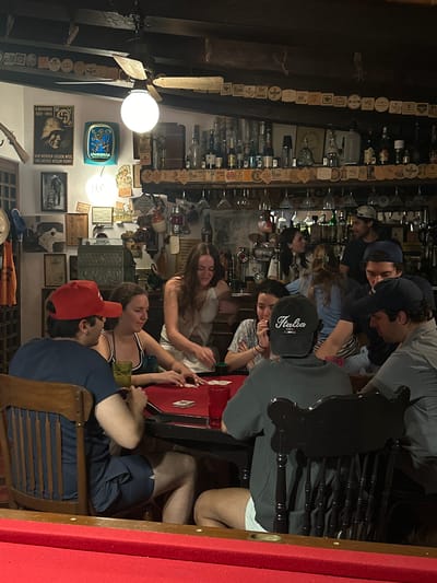 A group of approximately ten people, mostly young adults, are gathered around a red-topped table in what appears to be a pub or tavern with an extensive collection of beer coasters and memorabilia displayed on the walls. The room is dimly lit, illuminated by two prominent light fixtures: one being a round, exposed bulb and the other part of a ceiling fan fixture. Some people are wearing baseball caps and the mood seems casual. The overall environment is cluttered and evocative of an older establishment. Notably, the text in one of the posters on the wall appears to reference a "9 November 1923-1943" and "Adolf Hitler," and the term "Lowenbrau" appears on another wall decoration. The location appears to be Lequeitio, Mexico, though this is not evident from the image itself.