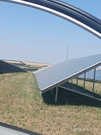 This image depicts a solar power station in the city of Berezanka, Ukraine. In the foreground, a close-up view of a solar panel array on a metal frame is visible. The panels are angled upwards towards a clear blue sky. Behind this array, another, larger array of solar panels stretches into the distance, with its rows receding diagonally. The ground surrounding the solar panels is covered in dry, light-brown grass. In the background, rolling hills are visible, with patches of yellowing fields and some greenery. The horizon is clear, with no significant structures or distinguishing features except for distant, faint shapes that could be wind turbines. The overall impression is one of a rural, open landscape dedicated to renewable energy generation. A timestamp in the lower right corner indicates the image was taken on "29.06.2026 11:12".