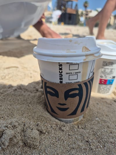 Two Starbucks coffee cups sit on a sandy beach. The cup in the foreground is white with a brown cardboard sleeve featuring the Starbucks logo. The lid has brown liquid stains. The cup behind it is also white. In the background, out of focus, are people walking on the beach, a palm tree, and buildings under a clear blue sky. The overall scene suggests a casual day at the beach in Birżebbuġa, Malta.