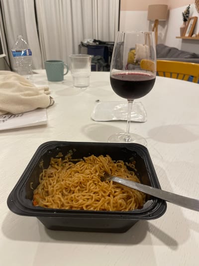 A close-up, high-angle shot depicts a still life on a white table. The primary objects are a partially-filled wine glass with red wine, a black plastic container filled with noodles, and a metal fork resting in the noodles. Other objects on the table include a water bottle, a small blue cup, a clear glass, a white cloth, and a white box. The background reveals a glimpse of an interior, possibly a residential space, with a bed frame and a lamp visible. Location cannot be determined from the image, and there is no basis to assume this is in El Tezal, Mexico. There are no people present in the image.