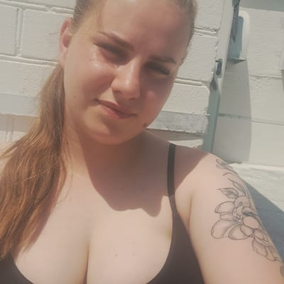 A young woman with long, auburn hair is lying on her back, possibly sunbathing. Her head is tilted upwards, and her eyes are closed, suggesting relaxation or sleep. She is wearing a dark-colored halter top or bikini top. A black tattoo of a floral design is visible on her upper right arm. The background appears to be a white, textured wall, possibly brick, illuminated by bright sunlight, casting shadows on the wall. The overall impression is one of a warm, sunny day. The image is cropped to focus on the woman's face, upper body, and arm, with the background providing context for an outdoor setting. No text is visible in the image. The location is stated as Bohdanivka, Ukraine.
