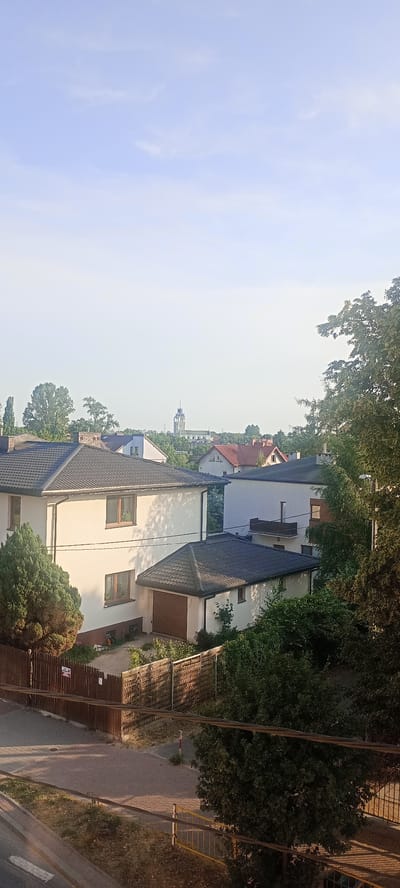 This is a medium-angle, vertical, outdoor shot of a suburban residential area in Ząbki, Poland. The scene is bathed in the soft, warm light of what appears to be early morning or late afternoon. The sky is a pale blue with a few wispy clouds, suggesting fair weather.

In the foreground, a two-story house with a dark tiled roof dominates the left side of the frame. It's painted a light cream color and features several windows with brown frames. Attached to the right side of this house is a smaller, single-story structure, possibly a garage, also with a dark tiled roof and a brown garage door. A portion of a wooden fence, partially obscured by lush green trees and bushes, runs along the front of the property. The fence casts long shadows on the paved walkway behind it. Further back, other houses with varying roof colors and architectural styles are visible, nestled amongst mature trees.

In the middle distance, a more distant view reveals a church steeple piercing the skyline, a characteristic landmark of many European towns. The presence of trees and the style of the houses strongly suggest a residential neighborhood in Poland.

The overall mood is peaceful and tranquil, evoking a sense of everyday suburban life. There are no people visible in the frame, and no discernible text is present. The focus is on the residential architecture and the natural elements, with the warm lighting creating a pleasant atmosphere.