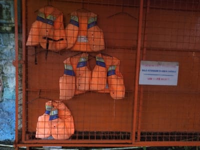 The scene presents an orange metal grid storage cage holding six bright orange life jackets, each accented with blue and light green/yellow sections near the shoulders, suspended by wire hangers. A white rectangular sign is affixed to the right side of the cage, featuring black text. It states "USE A LIFE JACKET" in English, and "SILA BERMAIN DI AREA DANAU" (Please play in the lake area) in Malay/Indonesian. To the left, a portion of a weathered, greenish-blue wall is visible. No individuals are present. The language displayed on the sign does not correspond to the specified location of Ciudad de San José, Costa Rica.
