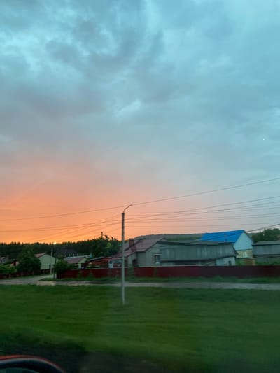 A rural landscape scene at dusk in Nurkeevo, Russia. The sky is a gradient of soft orange near the horizon, transitioning to a pale blue and then a darker, cloudy grey overhead. A dense line of trees forms a dark silhouette against the horizon. In the midground, several residential buildings are visible, featuring various roof colors including red, blue, and grey. A dark red fence runs along the front of some of these properties. A utility pole with wires is prominently placed in the right midground. The foreground consists of a grassy area, appearing slightly damp, suggesting recent rain. The image seems to be taken from a moving vehicle, as indicated by a blurred car interior visible in the bottom left corner.