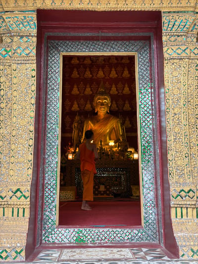 The image shows a monk standing barefoot in front of a large, golden Buddha statue inside a temple. The monk is wearing an orange robe and has his back to the camera. The Buddha statue is seated and is adorned with gold fabric. Behind the statue, there is a backdrop with a pattern of smaller Buddha images. The temple interior is richly decorated with gold and colorful mosaic tiles. The entrance to the temple is framed by ornate, gilded carvings and mosaic patterns, suggesting a significant religious or cultural site in Choumkhong, Laos. The floor inside the temple appears to be covered in a red carpet, while the floor outside the entrance has a patterned tile design.