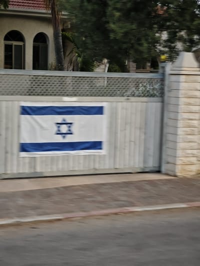 The image displays the Israeli flag displayed on a white, vertical wooden fence. The fence is part of a larger gate structure, with a stone pillar to the right. The background shows a building with arched windows and a tiled roof, partially obscured by trees. The scene is captured from a moving vehicle, indicated by motion blur in the foreground and midground. The location is identified as Yarchiv, Israel.