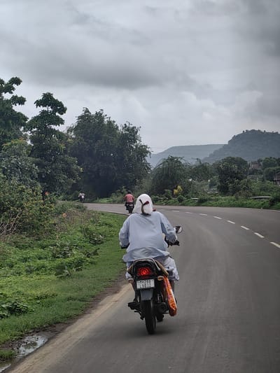 The image shows a person riding a motorcycle on a road in Rajur, India. The rider is wearing a light blue garment and a white head covering. The motorcycle has a license plate that reads "MH28B L9032". The road is paved and has white dashed lines indicating lanes. On the left side of the road, there is a grassy area with lush green vegetation and trees. In the background, there are rolling hills covered with trees, and the sky is overcast. Another motorcycle with a rider in a red shirt is visible further up the road. The scene appears to be a rural or semi-rural setting.