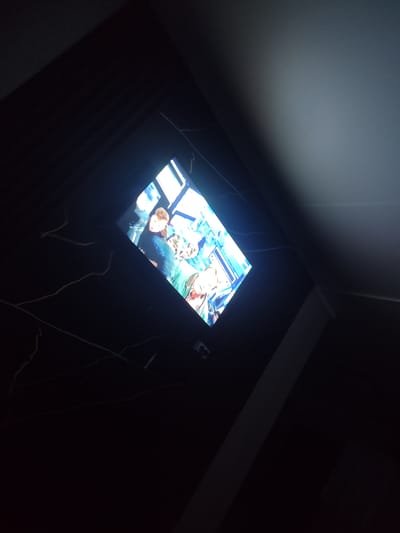 The image captures a close-up, angled view of a television screen displaying a group of people. The screen is illuminated with bright colors, predominantly blue and white, against a dark background. The people on the screen appear to be engaged in some activity, though their exact actions are unclear due to the image quality and angle. Some individuals have light-colored hair and are wearing clothing in shades of blue and green. The surrounding environment is dark, with some thin, white wires visible in the foreground, extending from the left side of the frame towards the television. A bright light source is casting a shadow on the upper right portion of the image, suggesting the presence of a ceiling light or external light source. The overall scene suggests an indoor setting where a television is being watched. The specific location within Қосшағыл ауылдық округі, Kazakhstan, cannot be determined from the visual information provided.