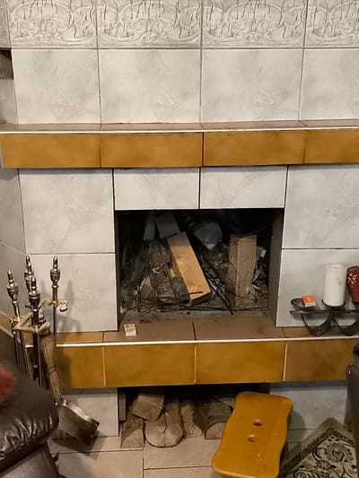 The image depicts an indoor fireplace with a tiled surround. The fireplace opening contains a pile of unburnt wood and debris. To the left of the fireplace, a set of brass fireplace tools, including a poker, brush, and shovel, are arranged on a stand. A dark brown leather armchair is partially visible in the foreground on the left. To the right of the fireplace, on a shelf, are a white candle, a red decorative object, and a small orange box. Below the fireplace, logs are stacked. A yellow wooden stool with two circular cutouts sits in front of the fireplace. A patterned rug is visible on the floor to the lower right. The environment appears to be a domestic interior. The location is identified as Žiaukos, Lithuania.