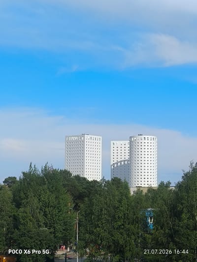 In Kogalym, Russia, a daytime cityscape features two modern, white, multi-story buildings with a grid pattern of windows, rising above a dense canopy of green trees. The sky above is a clear blue with wispy white clouds. The lower portion of the image reveals a street scene with people and a fence, partially obscured by the trees. The bottom right corner displays a timestamp: "21.07.2026 16:44". The bottom left corner shows "POCO X6 Pro 5G", indicating the device used to capture the image.