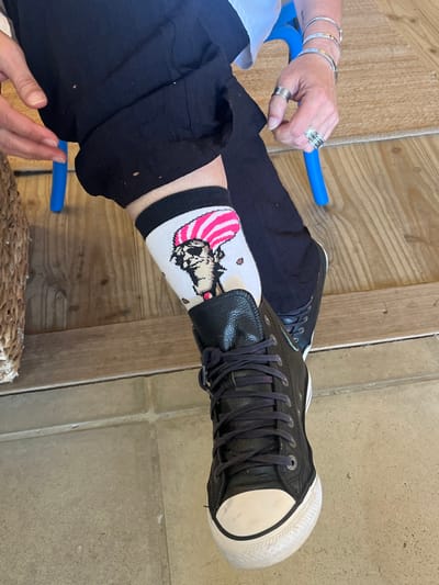 A person wearing black trousers and black leather high-top sneakers with white toes and white soles. On the left leg, the person has on a white ankle sock decorated with the image of a cartoon character with a pink-and-white striped hat. On the person's left hand are three rings and three silver bracelets. The person is sitting on a surface made of light-colored wood planks and next to a gray tile surface. A blue object is also visible.