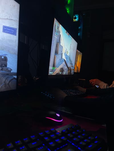 Inside a dimly lit gaming establishment in Karmana, Uzbekistan, multiple computer workstations are visible. The foreground features a dark desk with two keyboards, both exhibiting blue LED backlighting. A black gaming mouse, identified as Logitech G with pink and purple LED illumination, rests on a red surface in front of the keyboards.

To the left, a vertical monitor displays a digital street sign bearing "RUE PRINCIPALE" and "الشارع الرئيسي" (Main Street in French and Arabic), partially obscured by a dark object resembling machinery or a vehicle component at the bottom.

The central monitor prominently displays a first-person perspective video game, likely a shooter, depicting a character's hands gripping a knife in a desert-like urban combat environment with earthen walls and structures. Game interface elements, including a score or currency display of "$3350," are visible.

To the right of the central monitor, another monitor is partially visible, also displaying game content. Above the monitors, the illuminated internals of computer towers, featuring green and blue LED fan lighting, are visible in the dark background.

Two individuals are present. One individual's hands are positioned over a keyboard in the mid-ground, indicating active engagement with the central computer. To the far right, a second individual's hands are visible, holding a white smartphone, positioned near another keyboard. The overall scene is characterized by ambient light primarily emanating from the electronic displays and peripherals.