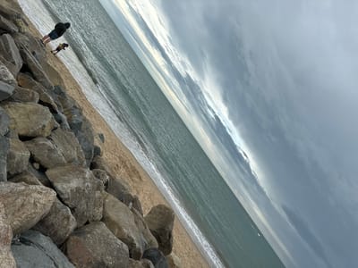 The image depicts a view of the sea from a rocky shoreline in Courtown, Ireland. Two individuals are visible: an adult wearing a dark hooded jacket and shorts, standing on the rocks, and a smaller figure, possibly a child, in colorful clothing, walking on the sandy beach. The sea is calm with gentle waves breaking on the shore. The sky is overcast with dark clouds, suggesting potential rain or a stormy weather front. The foreground is dominated by large, grey rocks forming a breakwater, with a sandy beach stretching alongside it. The overall scene conveys a sense of coastal natural beauty under a dramatic sky.