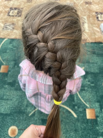 A top-down view shows a child's head with brown hair styled in a French braid. A yellow elastic band secures the end of the braid. The child wears a pink and gray plaid dress or shirt. The background features a green patterned carpet and a beige patterned surface. The location of Verkh-Katunskoye, Russia, is not verifiable from the image.