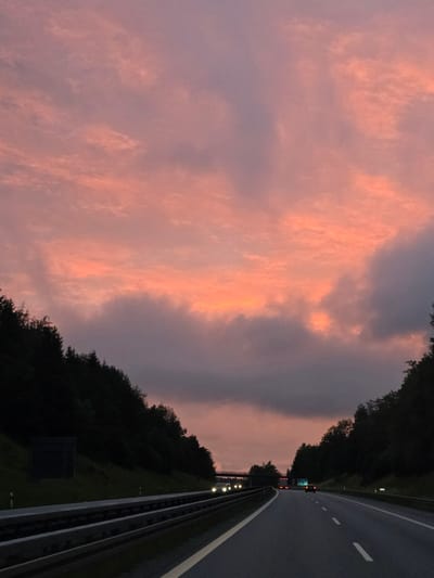 An exterior, eye-level, medium shot captures a highway in Niederwinkling, Germany, at dusk. The road is paved and appears to be a multi-lane highway with guardrails and white lane markings. Dark green trees line both sides of the road, forming dense forests that ascend the slopes. The sky is filled with dramatic clouds, painted in hues of pink, orange, and grey, indicating either sunrise or sunset. Several vehicles, including cars and trucks, are visible on the highway, their headlights on as the light fades. The overall scene conveys a sense of travel and the transition from day to night.
