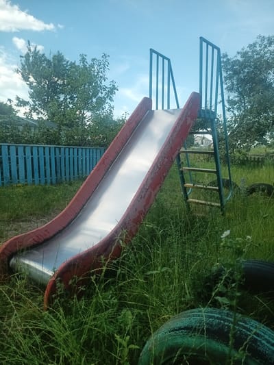 This media file shows a red and silver children's slide in a grassy outdoor setting. The slide has a metal surface and red, weathered edges. A blue metal ladder with wooden rungs leads up to the slide. The area around the slide is overgrown with tall green grass, and some old tires are visible in the foreground. In the background, there is a blue wooden fence and several trees. The sky is blue with scattered white clouds, suggesting it is daytime, likely late spring or summer. The overall impression is one of a slightly neglected or rustic playground. There is no visible text in the image. The setting appears to be a rural or suburban backyard in Velyka Dymerka, Ukraine. No people or specific activities are depicted, but the slide suggests a place for children's recreation. The weather appears pleasant and sunny.