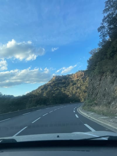 The scene depicts a mountain road in Les Cluses, France, viewed from the interior of a vehicle. A dark asphalt road with white dashed lane markings curves to the left in the foreground. A solid white line delineates the right edge of the road, alongside a concrete curb and a steep, rocky embankment densely covered with green trees and shrubs. Exposed reddish-brown rock formations are visible on the right hillside. To the left of the road, a concrete barrier is present, beyond which the terrain slopes downward, also covered in dense foliage. The background features a large, heavily vegetated mountain range with visible rock outcroppings. The sky is bright blue with scattered white cumulus clouds. The entire scene is well-lit by natural sunlight. No individuals, specific actions, or interactions are visible.