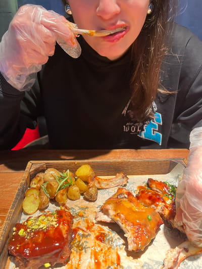 An indoor, close-up, high-angle shot depicts an individual consuming a meal at a wooden table within what appears to be a restaurant setting in Родниковское сельское поселение, Ukraine. The person's face is partially visible from the nose downwards, revealing dark hair and red lipstick. They are wearing a black hooded sweatshirt with a visible light blue and white graphic that includes "WOLF" on the right chest area. Both hands are covered with clear, disposable plastic gloves. The left gloved hand is holding a cleaned bone from a piece of meat, which is positioned near the person's mouth, indicating active eating.
The meal is served on a wooden tray lined with parchment paper, placed on the wooden table. The tray contains several pieces of roasted meat, likely ribs, in various stages of consumption, along with roasted baby potatoes garnished with fresh rosemary sprigs. One prominent piece of meat is coated in a thick, reddish-brown sauce and topped with crushed green pistachios and orange zest. Other pieces of meat are covered in a glossy, orange-brown glaze, some adorned with chopped green herbs. Extensive sauce residue and scattered bones are present on the parchment paper, indicating a significant portion of the meal has been consumed. The background is dark and out of focus, with a hint of a red object in the lower left, consistent with restaurant seating.