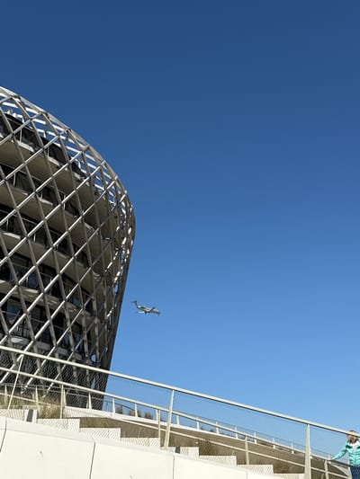 The image presents a low-angle architectural shot set against a brilliant, clear blue sky, indicating bright daylight conditions. On the left, a prominent modern building features a complex, grid-like facade composed of geometric, possibly diamond-shaped, patterns. This structure appears to be made of light-colored metal or concrete, with darker, recessed window panels. Its upper section exhibits a gentle curve, leading towards the center of the frame.
To the right, another contemporary building segment is visible, characterized by a smooth, light-toned wall and a distinctive metal mesh railing running diagonally upwards. At the extreme top right corner, a person is partially visible, looking down over this railing. They are wearing a light blue and white patterned top.
Suspended in the air between the two buildings, against the expanse of the blue sky, is a small, light-colored object resembling a drone or a model airplane, captured mid-flight. The strong sunlight creates clear shadows, emphasizing the architectural textures. The scene suggests a modern urban or developed environment, consistent with the location context of Middelkerke, Belgium. There is no visible text in the image.