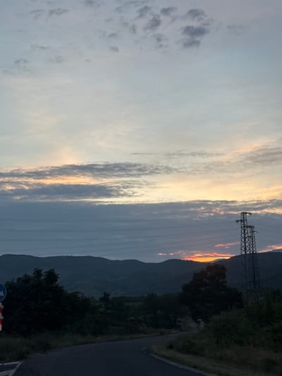 The image captures a landscape scene in Binkos, Bulgaria, at either sunrise or sunset, characterized by a dramatic sky and silhouetted terrain. The sky dominates the upper and central portions, featuring elongated clouds illuminated by warm orange and yellow light near the horizon, fading into lighter grey and pale blue further up. A striking band of bright orange light is visible peeking over the top of dark, silhouetted mountains or large hills in the mid-ground on the right.
A prominent electricity pylon stands on the right side of the image, its dark, latticed structure outlined against the sky, with multiple power lines extending horizontally across the mid-ground. Below the mountains, dense, dark green trees and vegetation line what appears to be a road or path running along the right side of the frame. In the bottom right corner, a partial view of a blue circular road sign and red and white striped barriers or cones indicates the presence of a road or construction area. There are no people visible, and no specific activities are taking place; the scene is a static depiction of a natural landscape infused with elements of human infrastructure. No legible text is visible.
