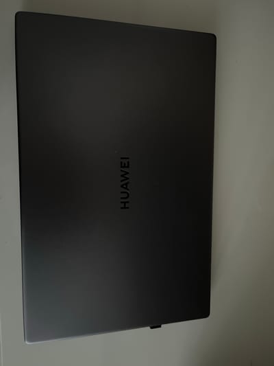 A dark gray Huawei laptop is closed and lying flat on a light-colored, smooth surface. The Huawei logo is embossed in the center of the laptop's lid. The scene is brightly lit from the left, casting a shadow to the right. The image was taken at an angle, with the laptop appearing slightly tilted. The background consists of a wall and a desk or table. There are no people or any other objects visible in the image. The location is the city of Maardu, Estonia.