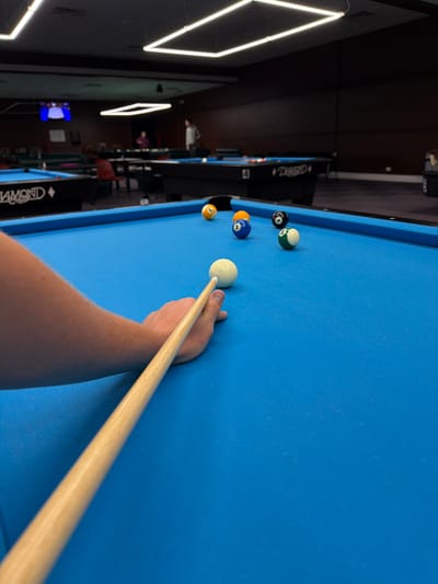 An over-the-shoulder, low-angle, close-up shot focuses on a person's arm and hand gripping a pool cue, positioned over a vibrant blue pool table. The cue is aimed at a white cue ball, which is slightly out of focus. Several numbered pool balls are scattered across the table in the mid-ground, including a yellow ball labeled "3", a blue ball labeled "10", a black ball labeled "8", and a green and white striped ball. In the background, a pool hall is visible with multiple pool tables, some with people playing, and ambient lighting from overhead fixtures. The location is a pool hall in Palatine, Illinois, United States of America.