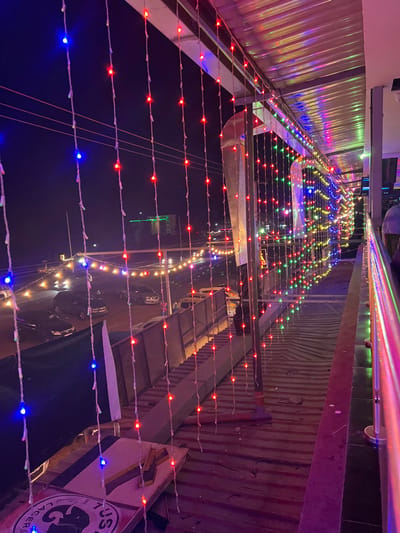 The image presents a vibrant night scene, looking down a narrow, illuminated outdoor space, possibly a street or a covered walkway, from an elevated perspective. Countless strings of multi-colored LED lights – predominantly red, blue, green, and yellow – are strung overhead, forming a dazzling canopy that stretches into the distance and creates a festive atmosphere.
On the left side of the pathway, a structure with corrugated metal siding is visible, reflecting some of the colorful lights. The right side features another building or wall, darker and also appearing to be constructed with corrugated metal. Below the dense network of lights, the dark ground surface reflects the illumination, suggesting it might be wet from recent rain or simply highly reflective. Blurry shapes of vehicles can be seen further down the path, indicating either parked cars or traffic.
The scene is clearly set at night, with the artificial string lights providing the primary source of illumination. No people are visible, but the setup implies an active commercial or entertainment area, perhaps a bustling street for dining or socializing. A prominent detail on the lower right side is a partially visible sign displaying the words "TUSKER" and "LAGER," along with part of an elephant logo. This identifies it as an advertisement for Tusker Lager, a well-known East African beer, consistent with the location context of Kimorori, Kenya.
