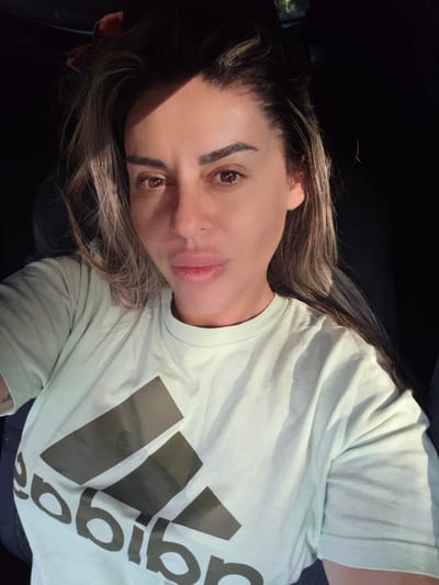A close-up, selfie-style photograph shows a woman with medium-toned skin, full lips, and brown eyes. She has dark brown hair with lighter highlights, styled loosely around her face. She is wearing a light green t-shirt with a large, dark gray Adidas logo on the front. The image appears to be taken inside a vehicle, with dark shadows indicating the interior of a car and bright sunlight casting dramatic highlights on her face and shirt. The background is predominantly dark and out of focus. The photograph was taken in the city of Ravnogor, Bulgaria.
