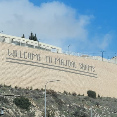 The image displays a large retaining wall on a hillside in Majdal Shams, Israel, under a partially cloudy blue sky. The wall, constructed of light-colored, possibly beige or light brown, textured blocks, features the dark gray pixelated text "WELCOME TO MAJDAL SHAMS" centrally placed. Two parallel dark gray horizontal lines run beneath the text. Above the retaining wall, a white building with a flat roof and white railings is visible, along with several dark green coniferous trees and multiple black streetlights with curved arms. The sky in the upper left quadrant shows a small, dark, airborne object. Below the retaining wall, the foreground consists of a rocky, sparsely vegetated hillside covered with dry grasses, small bushes, and some green foliage. Two black streetlights with curved arms extend upwards from the bottom of the frame, positioned on the hillside. No individuals or explicit actions are present.