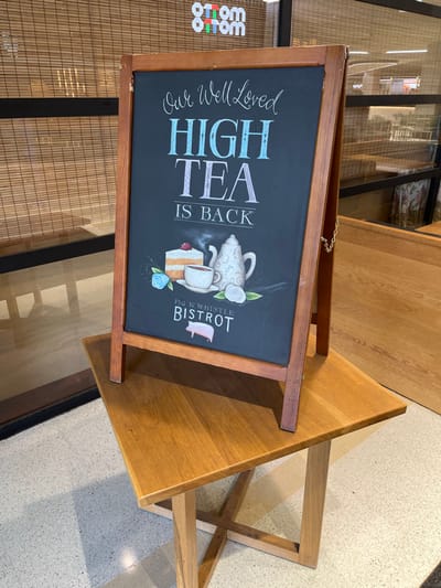 An A-frame chalkboard sign, framed in light brown wood, is prominently displayed indoors in what appears to be a commercial establishment, possibly a cafe or restaurant in Indooroopilly, Australia. The black chalkboard surface features a hand-lettered message in white, light blue, and light purple chalk. The main text reads, "Our Well Loved HIGH TEA IS BACK," with "HIGH TEA" in larger, decorative fonts. Below this, there's an illustration of a white teapot, a teacup with steam rising from it, and a slice of layered cake. Blue and white floral embellishments adorn the illustration. At the bottom right of the sign, "PIG N' WHISTLE BISTROT" is written, accompanied by a small pink pig logo.

The sign is placed on a light-colored tiled or concrete floor. To its left, a glass partition or door reflects a light brown, slatted screen or blind, and the white, reversed text "otom otom" is visible, likely indicating a nearby storefront. To the right of the sign, part of a wooden table with a light-colored top and light wooden legs is visible. The overall setting suggests a brightly lit, clean environment, likely aiming to attract customers with its high tea offering. No people are visible in the scene.