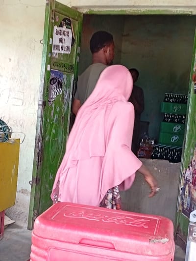 The image depicts a scene at the entrance of a modest shop or stall in Fanisau, Nigeria, during daylight. In the foreground, a woman in a pink hijab or layered garment stands with her back to the camera, facing into the interior. Beyond her, a man wearing a green t-shirt is visible inside the doorway, also with his back mostly to the viewer. A third person is partially obscured deeper within the shop.
A prominent red cooler with the "Coca-Cola" brand embossed on its lid is positioned in the immediate foreground. To the left, a portion of a yellow container is visible. Inside the shop, green crates stacked with bottled beverages are discernible on the right side. The overall activity suggests a customer or vendor interaction within a retail setting.
The shop entrance features a worn, green wooden doorframe and door. The door serves as an informal advertising space, displaying several pieces of text. Visible details include a phone number "8062597785," the name of a mobile payment service "OPAY," and the phrase "IMAMU Y'U SAN," likely a name or local language phrase. Another partial phone number, "070...", is also written on the door, along with smaller stickers featuring the "Coca-Cola" logo. The scene portrays a functional, local business environment.
