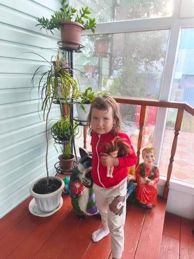 A young girl with curly brown hair stands on a wooden porch, holding a doll. She is wearing a red zippered jacket and beige pants with a teddy bear design on the leg. Next to her are two garden gnomes, one resembling a cat and the other a woman. In the background, several potted plants are arranged on tiered stands. The porch has a light blue, horizontally paneled wall to the left and a large window behind the plants and gnomes, looking out onto a yard with trees and buildings. The sun is shining, suggesting it is daytime, possibly late afternoon given the warm light. The setting appears to be a residential area in Burayevo, Russia, likely a house or dacha with a sunroom or enclosed porch. The girl looks directly at the camera with a neutral expression.