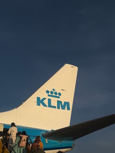 A KLM Boeing 737-800 aircraft is depicted during boarding at Stockholm-Arlanda Airport, Sweden. The aircraft's vertical stabilizer (tail fin) is prominently featured, white/cream in color, with the blue KLM logo – a crown symbol above the letters 'KLM' – visible on its side. Below the tail, a blue segment of the fuselage with the text '737-800' is partially visible. A section of the aircraft's dark underside of the right wing extends from the lower right foreground. A group of approximately six individuals is ascending bright yellow-orange boarding stairs, entering the aircraft's cabin. They are dressed in various casual attire; notable figures include a person in a light trench coat, another with a green outfit and a light brown backpack, and another with a dark brown backpack and an orange shirt. The background consists of a clear, deep blue sky, indicating an outdoor, daytime scene with direct sunlight illuminating the aircraft and people.