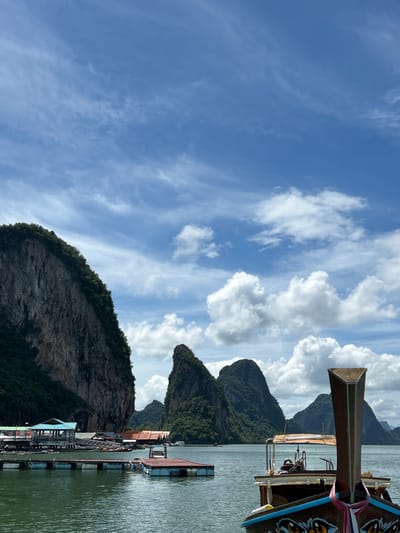 The image depicts a view of Ko Panyi, a village in Thailand known for its stilted houses and floating structures. In the foreground, the bow of a wooden longtail boat is visible, decorated with a colorful ribbon and intricate carvings. The boat is positioned on a body of calm water. In the mid-ground, a series of interconnected wooden pontoons and platforms extend into the water. On the left side of the image, a cluster of buildings with various colored roofs, including one with a turquoise roof, are situated on stilts over the water, forming part of the village. Behind the village, dramatic karst limestone formations rise steeply from the sea, covered in lush green vegetation. The sky above is a bright blue, with scattered white clouds, some wispy and others more voluminous, indicating a partly cloudy day. The overall scene is a picturesque representation of a coastal village in a tropical environment, characterized by water, boats, unique architecture, and striking natural geological features.