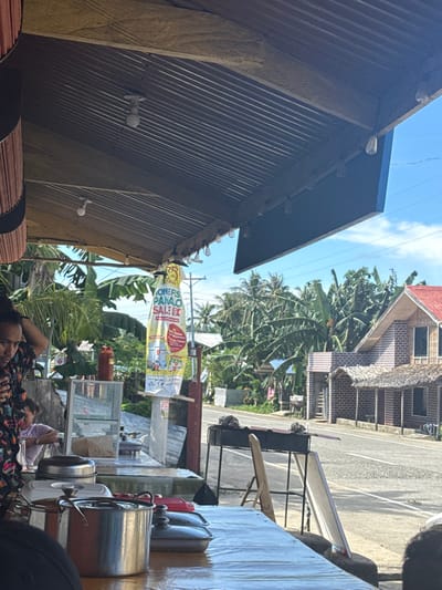The image depicts an outdoor or semi-outdoor eatery located alongside a road in Burgos, Philippines, on a clear, sunny day. In the immediate foreground, a wooden table with a plastic covering holds multiple stainless steel cooking pots and lidded serving trays. To the left, a person with dark, curly hair wearing a patterned shirt is partially visible, looking down and to the left, with their left hand resting on their head. Another person in a light-colored top is partially visible behind a glass display case on a counter further back. A red plastic bottle is also on this counter.

Above, the establishment's roof is constructed from corrugated metal sheeting, with multiple LED bulbs in white sockets and decorative string lights suspended from it. A tall, yellow and white banner with red text reading "ONERS PANAO SALE!" and "GET A CHANCE TO WIN..." hangs vertically from a wooden post.

Across a two-lane asphalt road in the midground, a black metal barbecue grill stand with items on top is visible, accompanied by a wooden chair. Further across the road, a distinct house with a red roof and brick-patterned exterior features several smaller, attached structures with thatched roofs. The background is dominated by dense tropical vegetation, including numerous banana and palm trees, under a bright blue sky. A utility pole with power lines is also visible.