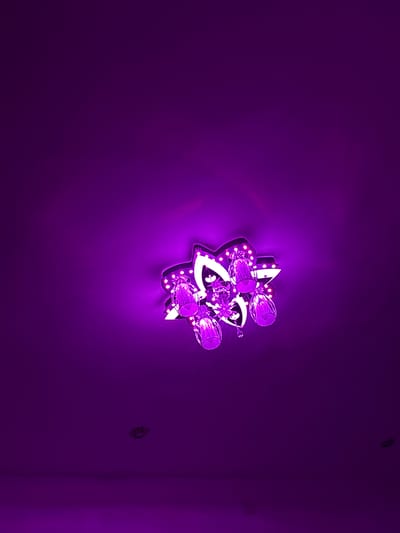 A vibrant purple wall is illuminated by a decorative, star-shaped light fixture centrally mounted. The fixture, characterized by a geometric design, features multiple small, round LED lights embedded along its edges, emitting a bright white-purple glow. It is adorned with clear, crystalline embellishments, including several cylindrical elements and smaller star-shaped accents that refract the light.
The intense illumination from the fixture creates a radiant purple halo on the immediate wall surface, fading into a deeper purple further away. The wall itself exhibits a subtle, granular texture. In the upper right corner of the frame, a small, dark, circular object, possibly a screw head or wall plug, is visible. Towards the lower right, a dark vertical line suggests a corner of a room or an adjacent wall, where a faint, small, dark rectangular object, possibly an electrical outlet or fixture, is present. The scene is indoors, presenting a distinct and modern aesthetic dominated by shades of purple. The context places this scene in Gorno Sahrane, Bulgaria. No text is visible in the image.