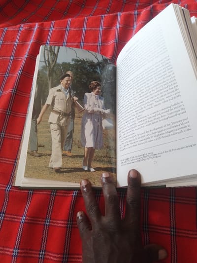 An open book lies on a red and black plaid fabric. The book's pages feature a photograph of a man and a woman standing in a grassy field. The man, wearing a safari-style shirt, pants, and hat, is holding a rifle. He is looking at the camera. The woman, wearing a light-colored dress, is also looking at the camera. Both figures appear to be in an outdoor setting. The book's text is partially visible on the right-hand side of the image.  The image appears to be a photograph taken from a book.  The coordinates (latitude: 40.688, longitude: 46.388) indicate the location is in the city of Ganja, Azerbaijan.