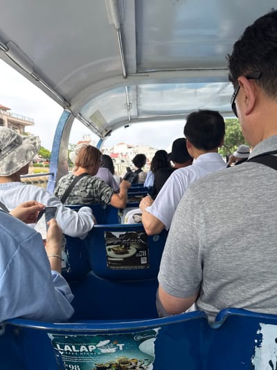 An eye-level, indoor shot of passengers on what appears to be a bus or tour vehicle. People are seated in rows, with the backs of the seats facing the camera. The seats are blue with advertisements, including food promotions. The passengers are dressed in casual attire, and some are looking at their phones. The bus has a curved roof with structural supports. The shot is taken at an angle, giving a distorted perspective.