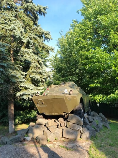 This is a photograph of a military vehicle, possibly an armored personnel carrier, positioned as a monument or memorial. It is painted olive drab and appears to be a static display. The vehicle is resting on a pile of large, rough-hewn rocks. Behind the vehicle, lush green trees with dense foliage are visible against a clear blue sky. To the left and foreground, a large coniferous tree with feathery, blue-green needles is prominent, its branches drooping. The setting appears to be an outdoor area, likely a park or memorial site.
There are no people visible in the image. Small red flowers are placed on the vehicle's exterior, suggesting a commemorative purpose. Some graffiti or markings are visible on the side of the vehicle. The lighting suggests it is daytime, with sunlight casting shadows on the rocks and grass. The overall atmosphere is solemn and reflective, fitting for a memorial.
The location context provided is Ivankiv, Ukraine. While no explicit text is easily legible on the vehicle, the context of a memorial monument in Ukraine could imply a connection to historical events or conflicts. The image is oriented such that the sky is on the left side, and the ground is on the right, indicating the photograph was taken with the camera rotated.