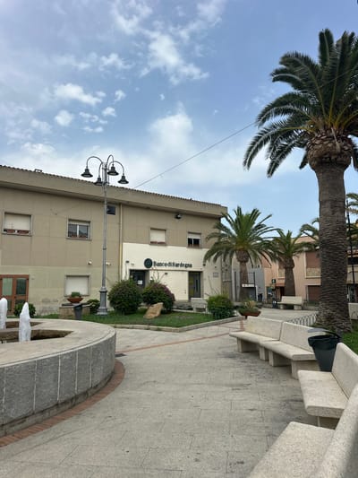 The image captures a view of a town square with a fountain in the foreground on the left. The fountain has multiple water jets. To the right of the fountain are several long, light-colored stone benches. Beyond the benches, there is a paved area and a green grassy area with bushes and a large rock. In the background, a building with windows is visible, featuring a sign that reads "Banco di Sardegna". Several palm trees are scattered throughout the scene. The sky is cloudy with patches of blue. The scene appears to be in Crabonaxa/Villasimius, Italy.