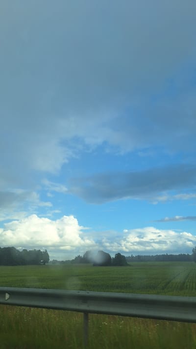 The image depicts a wide-angle view of a rural landscape in Glāžšķūnis, Latvia, under a dynamic sky. In the foreground, a weathered metal guardrail runs horizontally across the frame, its surface showing signs of exposure to the elements. Beyond the guardrail, a dense line of green grass and wildflowers stretches across the bottom of the image. The middle ground is dominated by a vast, flat field of green crops, likely grain or corn, stretching towards a distant tree line. The field is marked by subtle rows, suggesting agricultural cultivation. The far background features a dense, dark green forest that forms a natural horizon. The sky is a prominent feature, with a mix of bright blue sky interspersed with dramatic, volumetric clouds. These clouds range from brilliant white, fluffy cumulus formations to darker, more ominous grey storm clouds, suggesting a variable weather condition. There are no people or animals visible in the scene, and the overall impression is one of a serene, natural environment under a changing sky. The image appears to have been taken from a moving vehicle, as evidenced by the slight blur in the foreground foliage and the perspective.