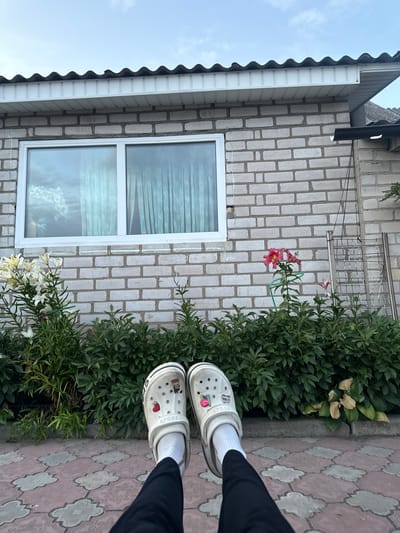 The image captures a low-angle view of a person's feet wearing white Crocs with various charms, white socks, and dark pants. The feet are positioned in front of a dense green hedge. Behind the hedge, a light-colored brick building is visible, featuring a white-framed window with sheer curtains. To the right of the window, there are two pink lilies. The roof of the building is corrugated and dark-colored. The ground in front of the hedge is paved with interlocking square tiles featuring a cloud-like pattern. The overall setting appears to be an outdoor area, possibly a backyard or garden, with elements suggesting a residential location within the Сароцкі сельскі Савет, Belarus. The sky is overcast with light clouds.
